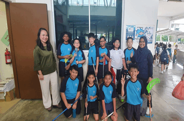 A group of children in blue jerseys holding floorball sticks pose with two adults inside a school building.